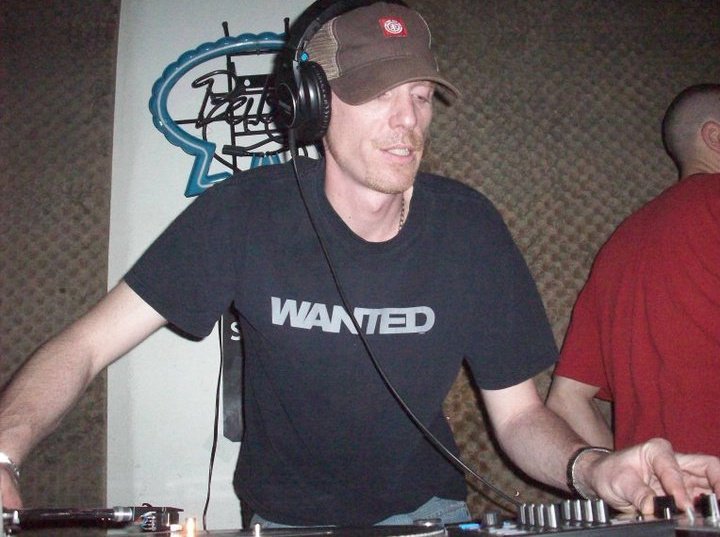 DJ Natural Nate® at the decks wearing a Wanted shirt and headphones
